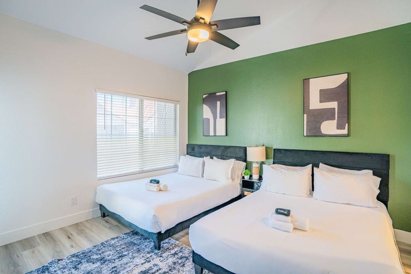 A hotel room with two beds, a ceiling fan, a window with blinds, and artwork on the green wall.