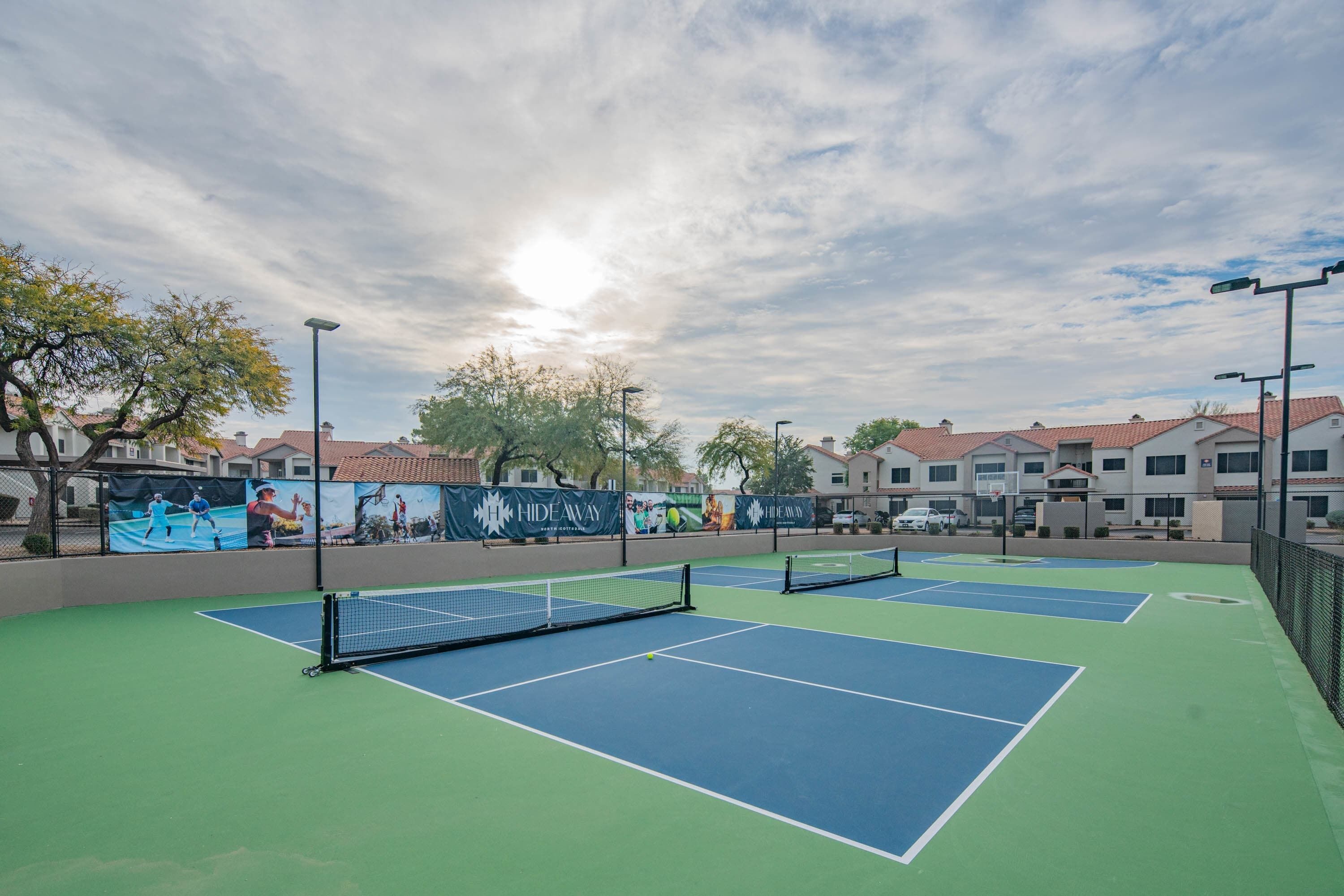 Un court de tennis extérieur doté de plusieurs terrains, entouré de bâtiments et d’arbres.