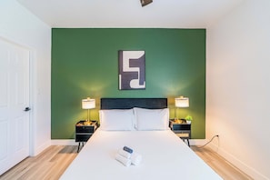 Quarto de hotel com uma cama grande, duas mesas de cabeceira, uma parede verde e uma obra de arte abstrata moderna.