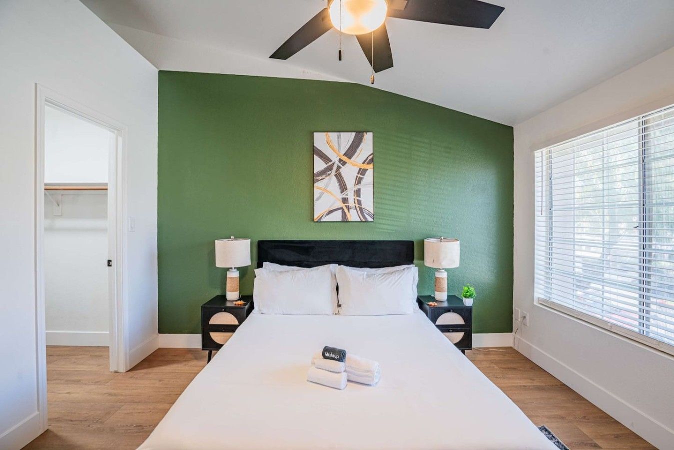 A bedroom with a bed, two bedside tables, a ceiling fan, a window with blinds, and a green accent wall.