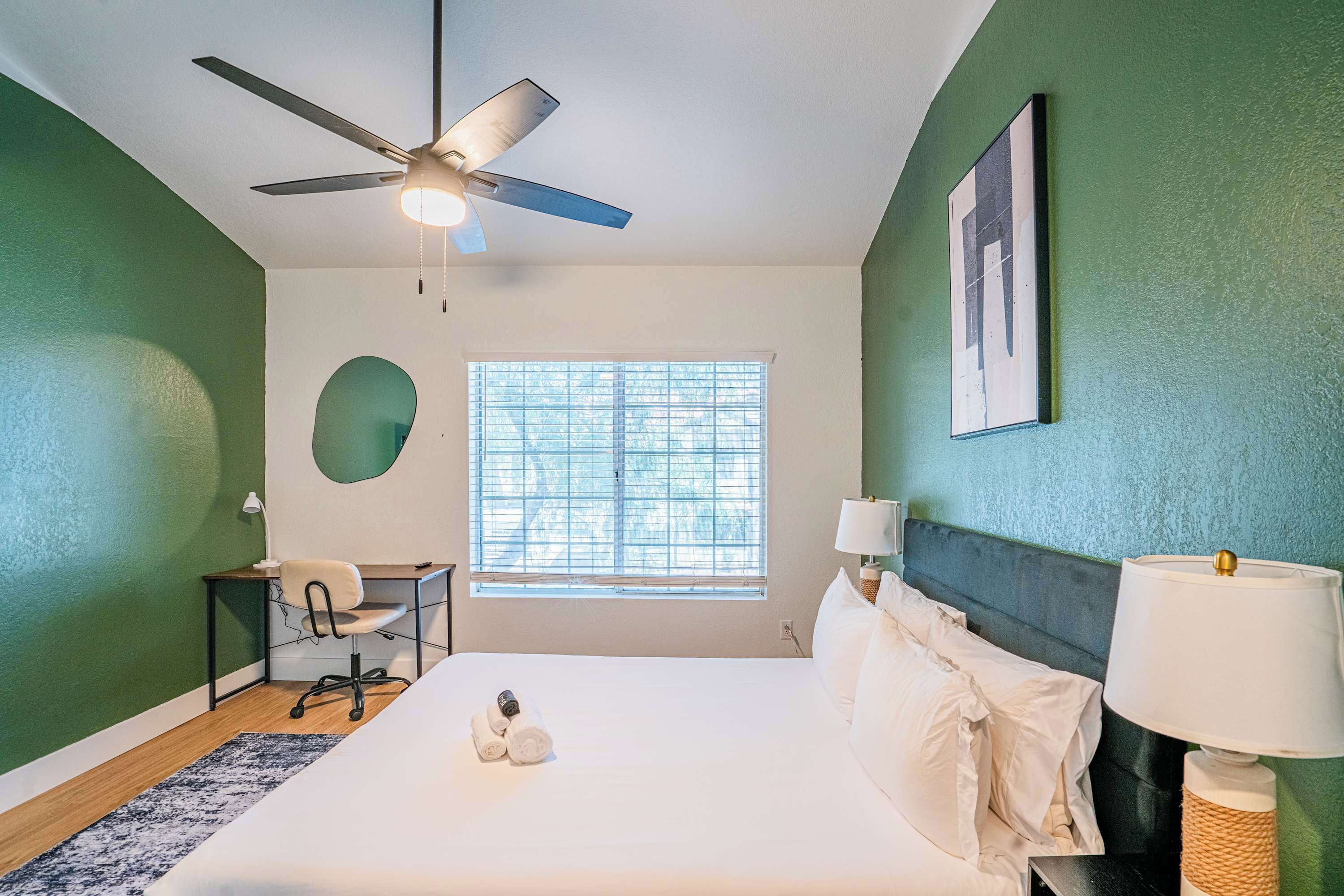 Une chambre d’hôtel équipée d’un lit, d’un bureau, d’une chaise, d’un ventilateur de plafond, d’une fenêtre avec stores et d’une lampe fixée au mur.