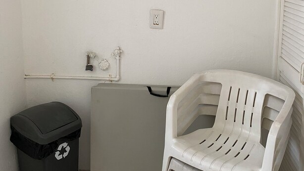 A small, enclosed space with a white wall, a black trash bin with a recycling symbol, a grey appliance, and a white plastic chair. - Monthly Beach House Rental in Manzanillo, Mexico – Perfect for Snowbirds (Manzanillo)