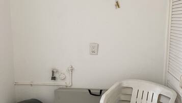 Un petit espace clos avec un mur blanc, une poubelle noire ornée du symbole du recyclage, un appareil électroménager gris et une chaise en plastique blanche.