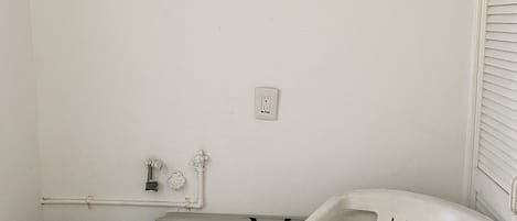 A small, enclosed space with a white wall, a black trash bin with a recycling symbol, a grey appliance, and a white plastic chair.