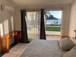 Ein Hotelzimmer mit Bett, Schreibtisch und Blick auf einen Strand mit einer Reetdach-Sonnenschirm.