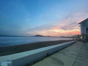 A coastal view with a beach, a paved walkway, and a building with a gated entrance.