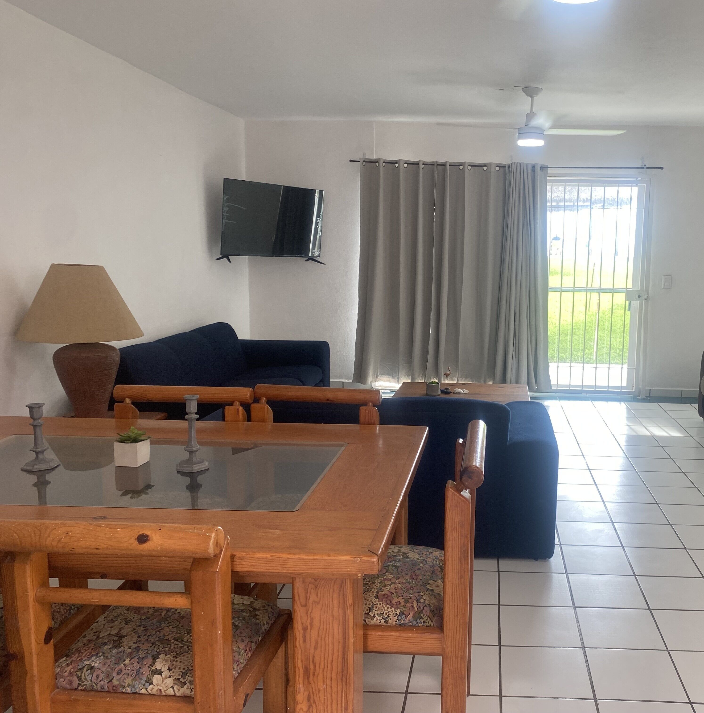 Una sala de estar con televisor montado en la pared, un sofá, una mesa de comedor con sillas y vistas a través de una ventana con cortinas.
