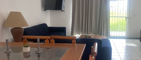 Una sala de estar con televisor montado en la pared, un sofá, una mesa de comedor con sillas y vistas a través de una ventana con cortinas.