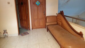 Una habitación con un sofá marrón, una puerta de madera con un ventanal en forma de rombo y suelo de baldosas.