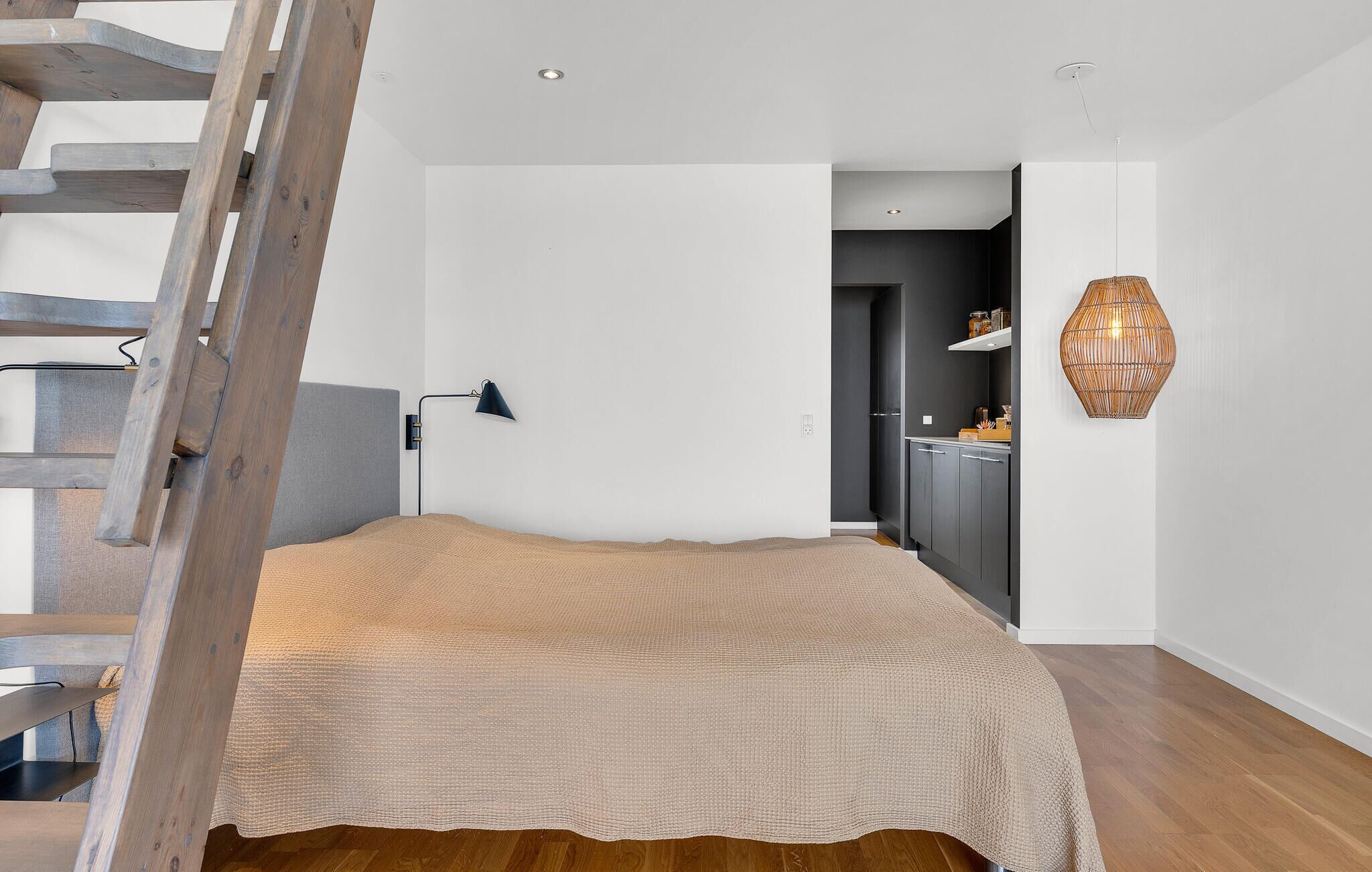 Un dormitorio con una escalera de madera, una cama con una manta texturizada, una lámpara fijada a la pared y una cocina visible a través de una puerta abierta.