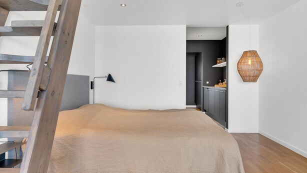 Un dormitori amb una escala de fusta, un llit amb llençols amb textura, una làmpada penjada a la paret i una zona de cuina visible a través d'una porta oberta.