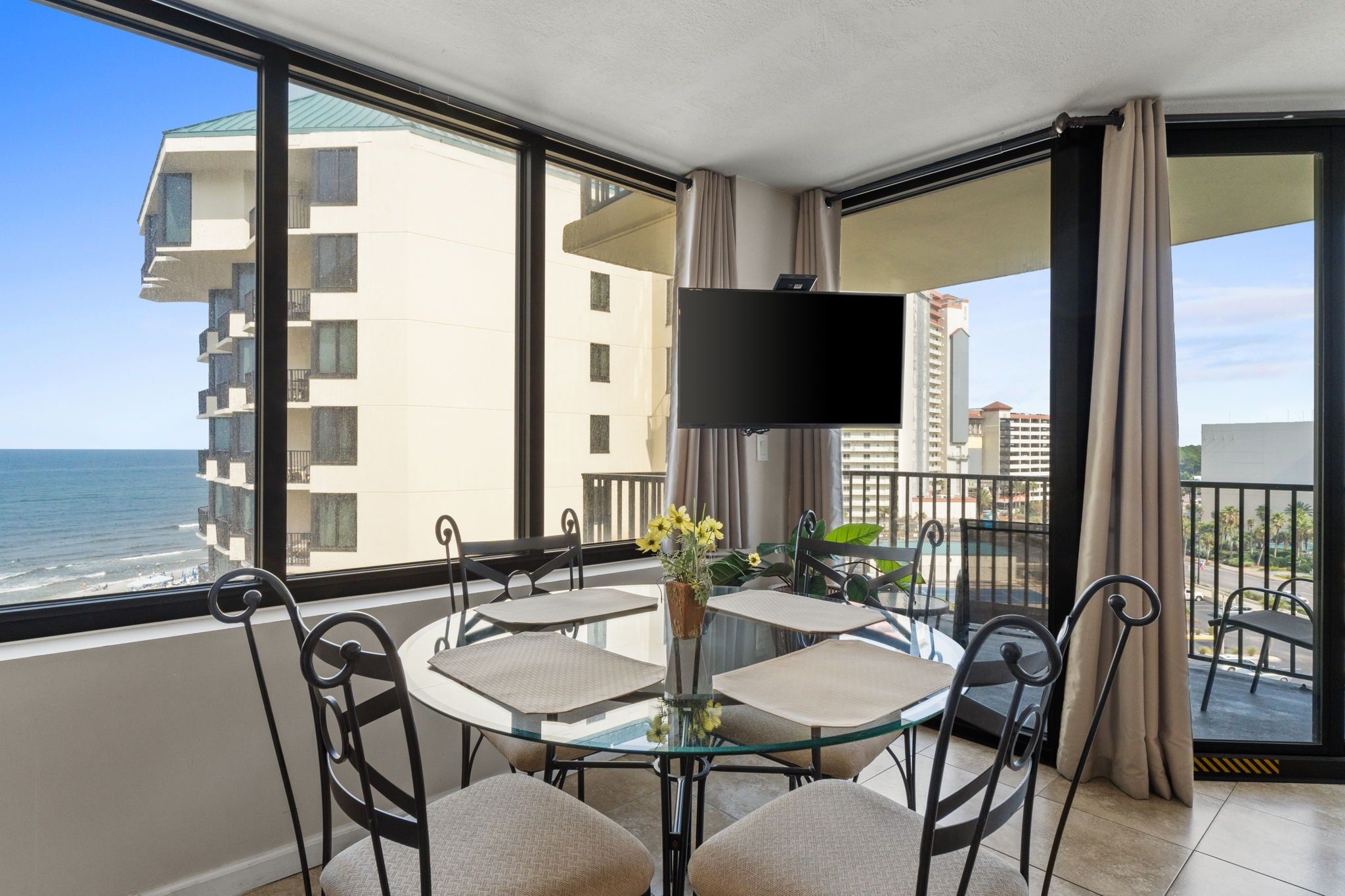 A balcony with a glass table and chairs, a flat-screen TV, and a view of the ocean.