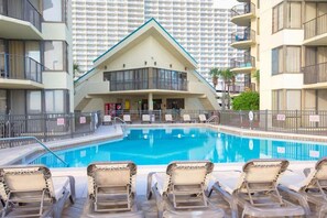 A swimming pool with lounge chairs, a building with a triangular roof, and a high-rise background. - Renovated Beachfront, Gulf Views & 3 Pools | Sunbird 912E (Panama City)