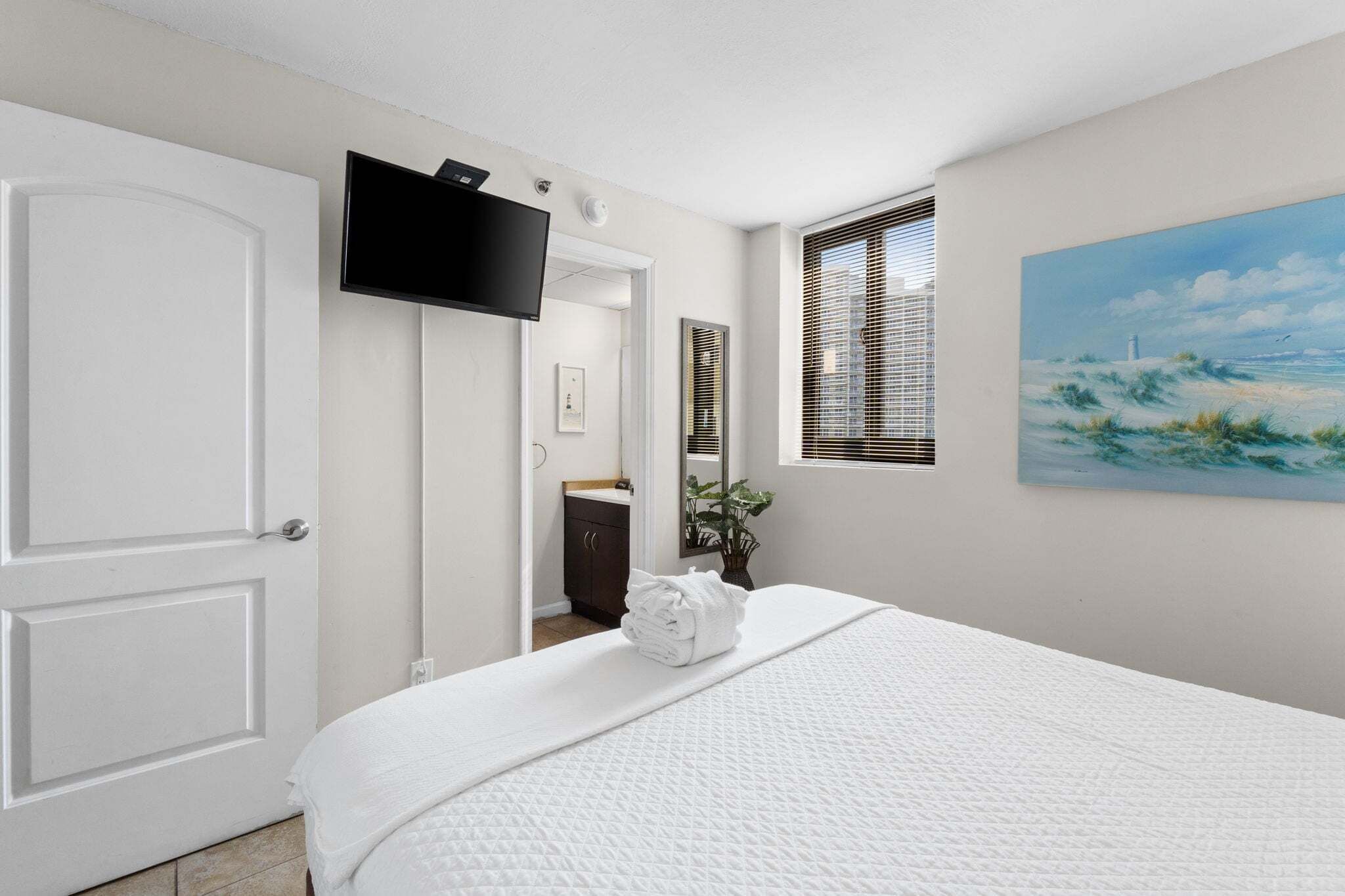 A hotel room with a bed, a television, a painting, and a window with blinds.