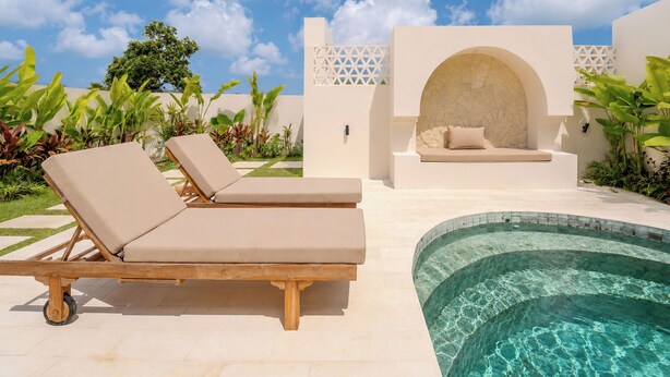 Un área junto a la piscina con sillones de descanso, una zona de estar con pared de piedra y abundante vegetación.