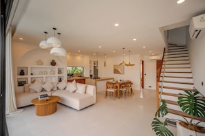 A modern living room with a sofa, coffee table, and dining area with wooden chairs. - Aintrazy Villa by Betterplace (Unggasan)
