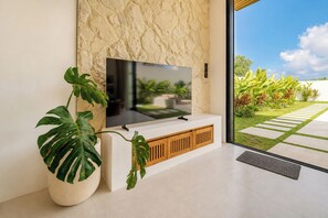 Un salón moderno con un televisor de pantalla plana montado en un mueble blanco, una planta grande en maceta y vistas a un jardín cuidado a través de un ventanal.