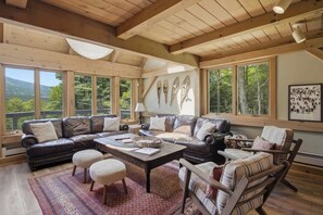 A spacious living room with a large window, leather sofas, a coffee table, and a fireplace. - Round Hill Retreat | Epic Stratton Views w Hot Tub (Winhall)
