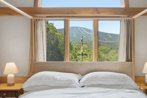 A bedroom with a bed, two bedside tables, and a window with a view of a mountain and trees. - Round Hill Retreat | Epic Stratton Views w Hot Tub (Winhall)