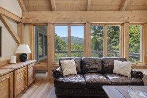 A wooden cabin interior with a leather sofa, a lamp, and a view of a mountain through large windows. - Round Hill Retreat | Epic Stratton Views w Hot Tub (Winhall)