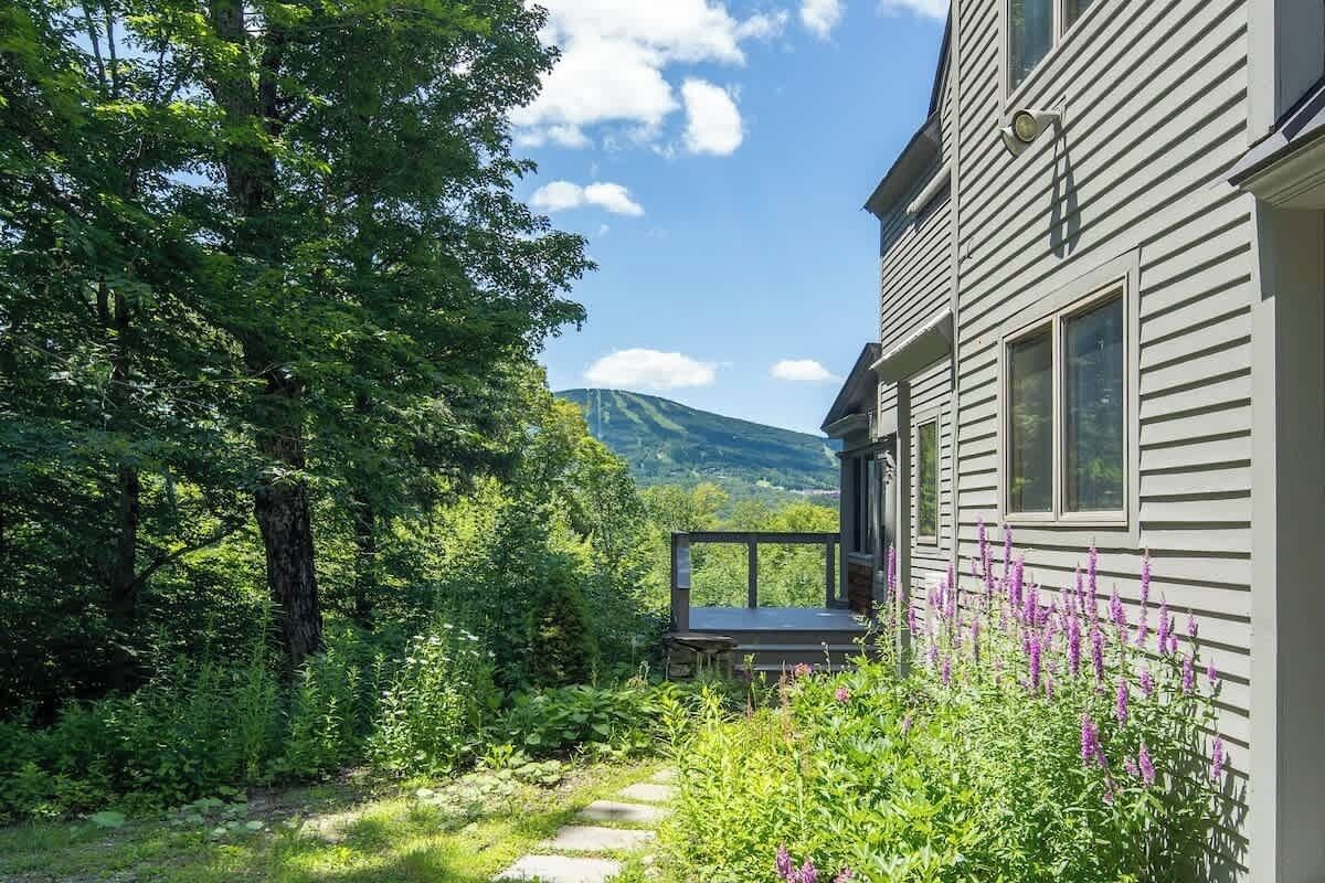 A house with a deck, surrounded by lush greenery and a mountain in the background.