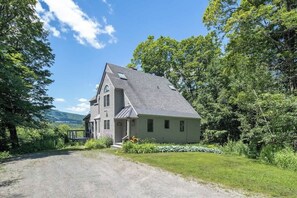 A two-story house with a sloped roof, a front porch, and a driveway leading to the entrance. - Round Hill Retreat | Epic Stratton Views w Hot Tub (Winhall)
