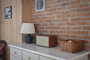 A room with a brick wall, a white dresser, a lamp, a vintage radio, and a wicker basket. - Round Hill Retreat | Epic Stratton Views w Hot Tub (Winhall)