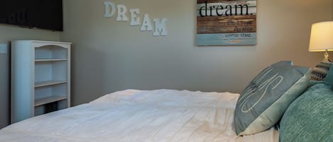 Une chambre à coucher comprenant un lit, un téléviseur fixé au mur, une étagère et des œuvres d’art décoratives aux messages inspirants.