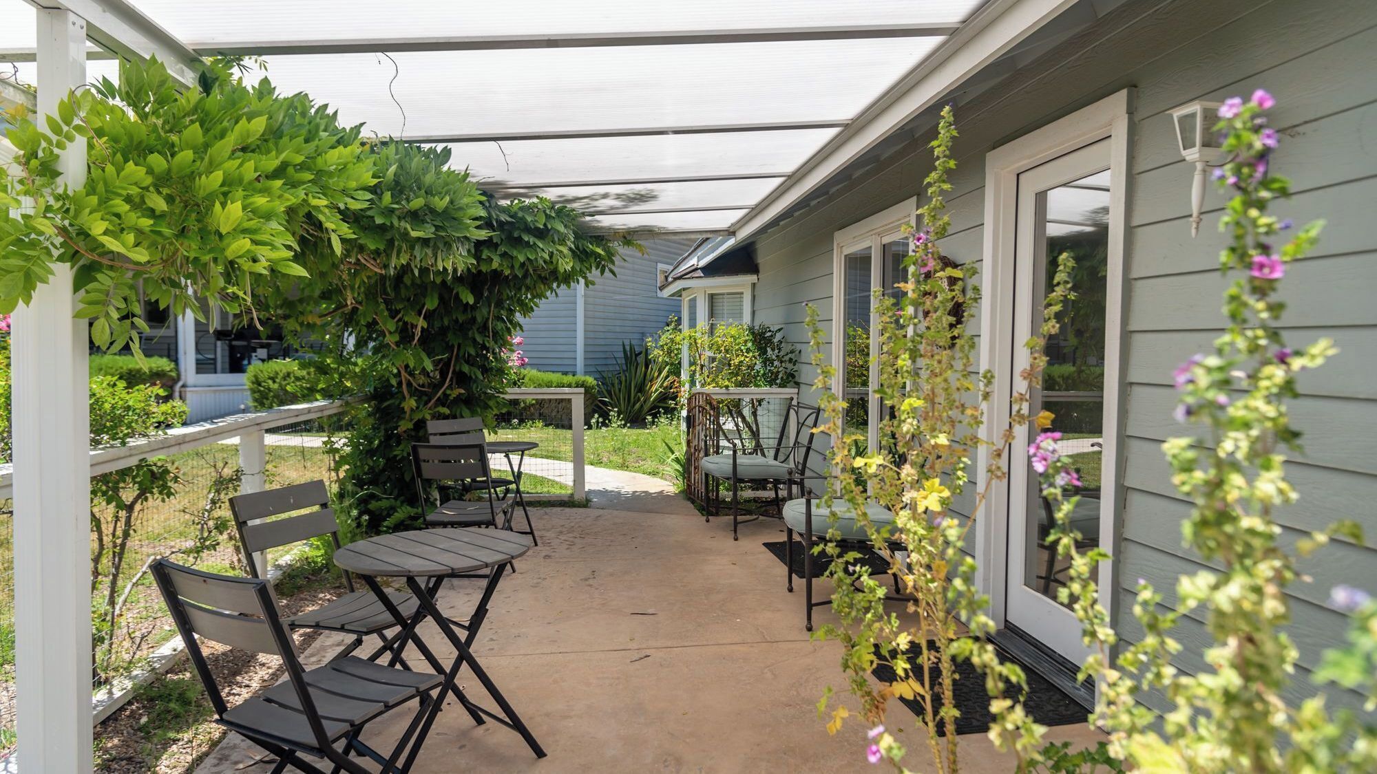 Un patio couvert avec une table et des chaises, entouré de verdure et d’une maison.