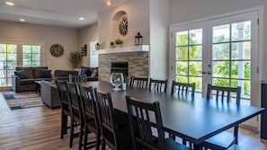 Ein großzügiger Essbereich mit einem großen Tisch und Stühlen, einem Kamin und Blick auf Grün durch große Fenster.