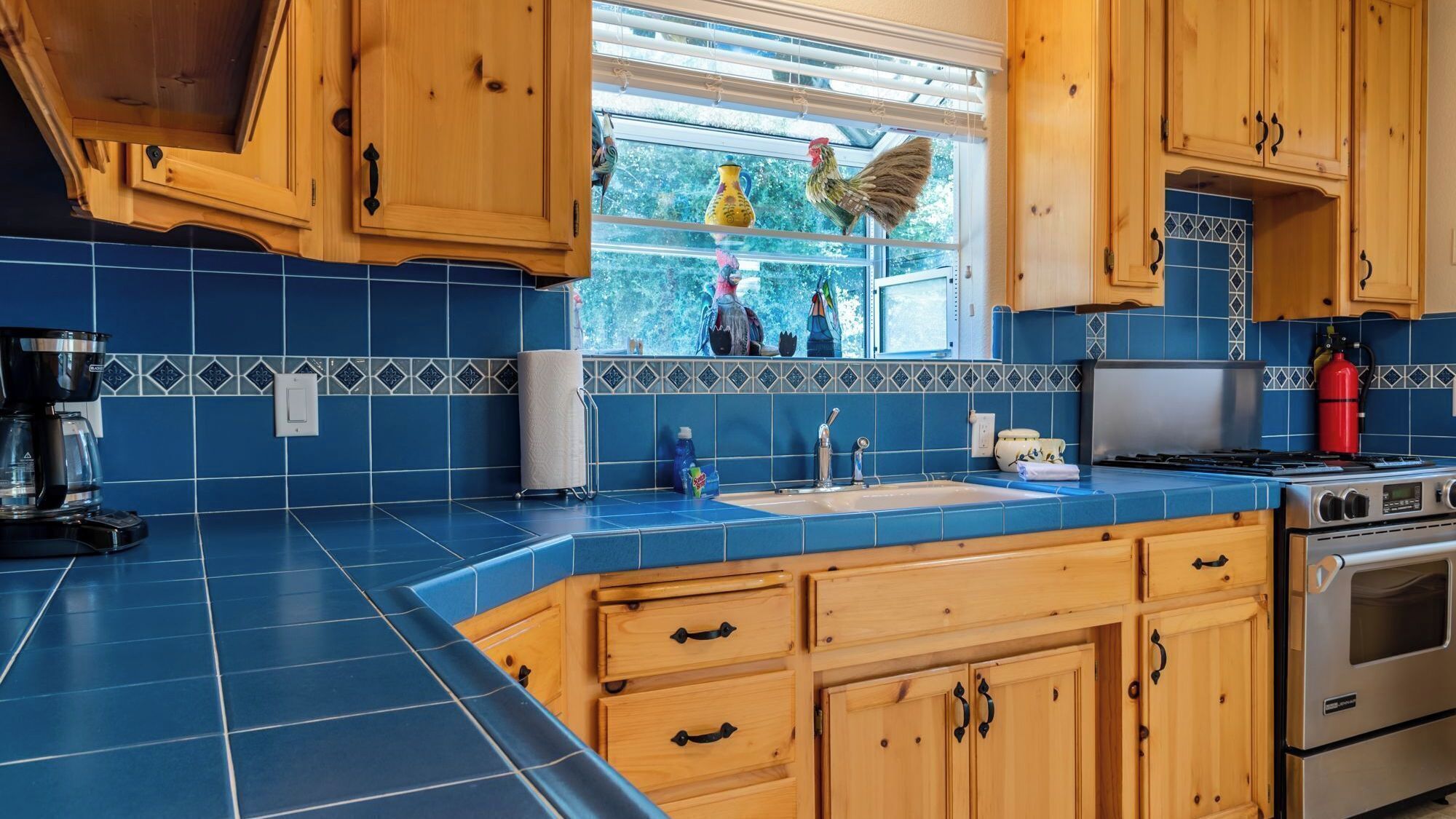 A kitchen with blue tiles, wooden cabinets, a stainless steel stove, and a window with decorative items.