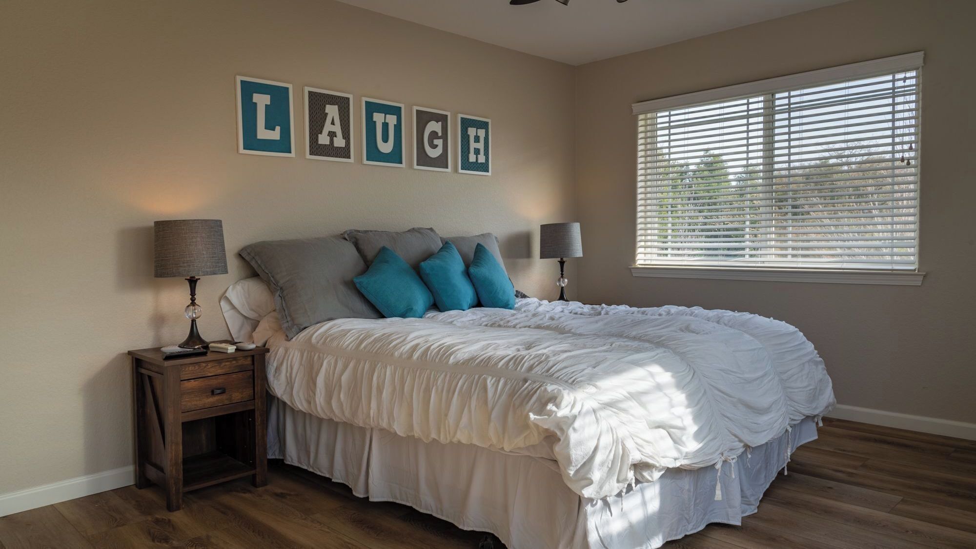 A bedroom with a bed, two bedside tables, a window with blinds, and a wall decoration spelling 'LAUGH'.