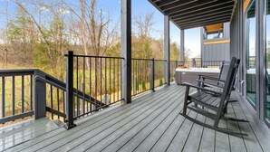 A wooden deck with a black railing, a bench, and a view of trees and a building. - CampingThemed/Pool/HotTub/GameRoom/FirePit/Sleep16 (Sevierville)