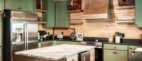 Una cocina con armarios verdes, electrodomésticos de acero inoxidable, una isla de mármol y una lámpara colgante.