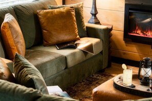 A cozy living room with a green sofa, leather cushions, a fireplace, and a small table with a candle and a glass. - CampingThemed/Pool/HotTub/GameRoom/FirePit/Sleep16 (Sevierville)
