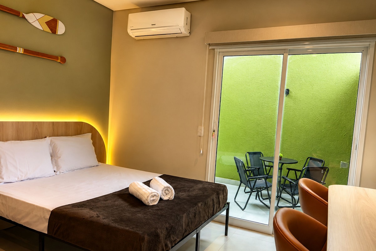 A hotel room with a bed, a sliding glass door to a balcony, a table and chairs outside, and an air conditioning unit on the wall.