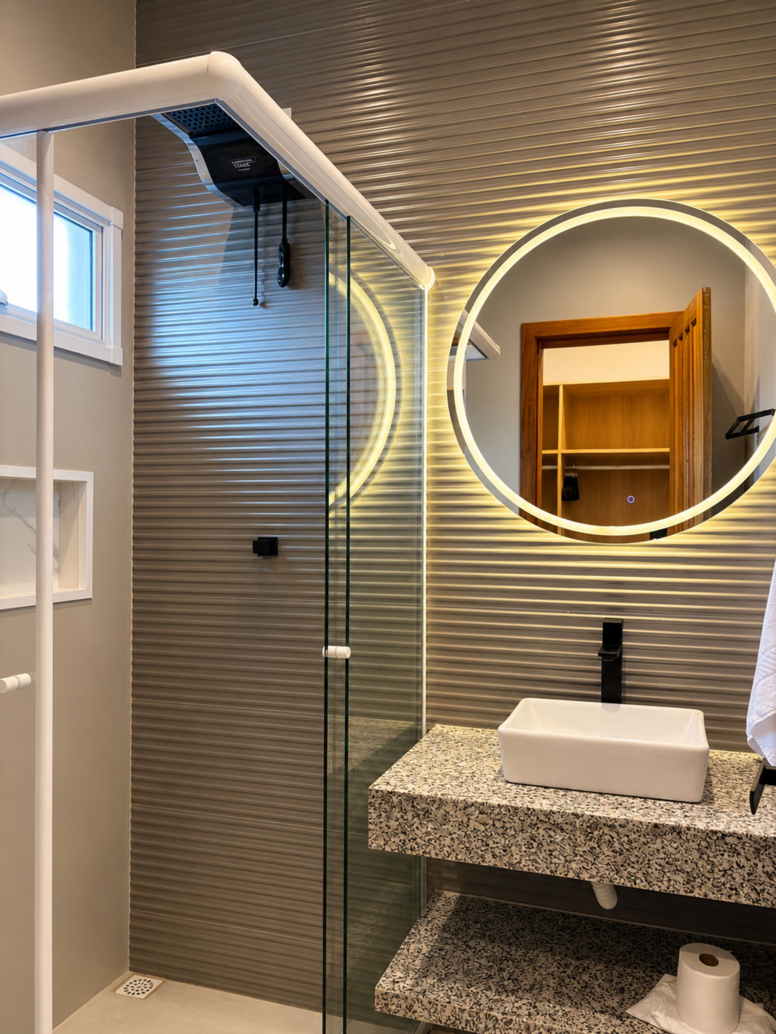 Une salle de bain moderne avec une douche à l’italienne en verre, un grand miroir rond encadré en bois et un plan de toilette en granit avec un lavabo.