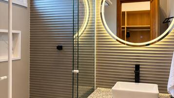 Une salle de bain moderne avec une douche à l’italienne en verre, un grand miroir rond encadré en bois et un plan de toilette en granit avec un lavabo.
