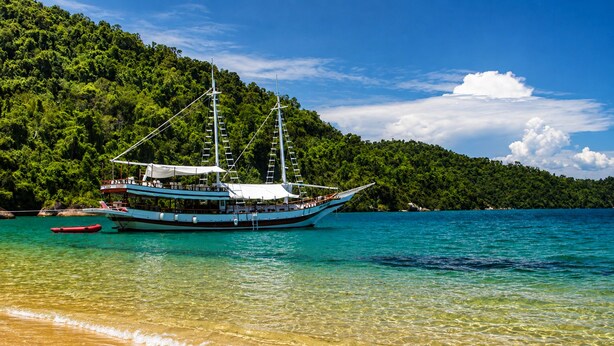 A boat anchored near a beach with clear turquoise water and a forested hill in the background. - Sabará Flats (Paraty)