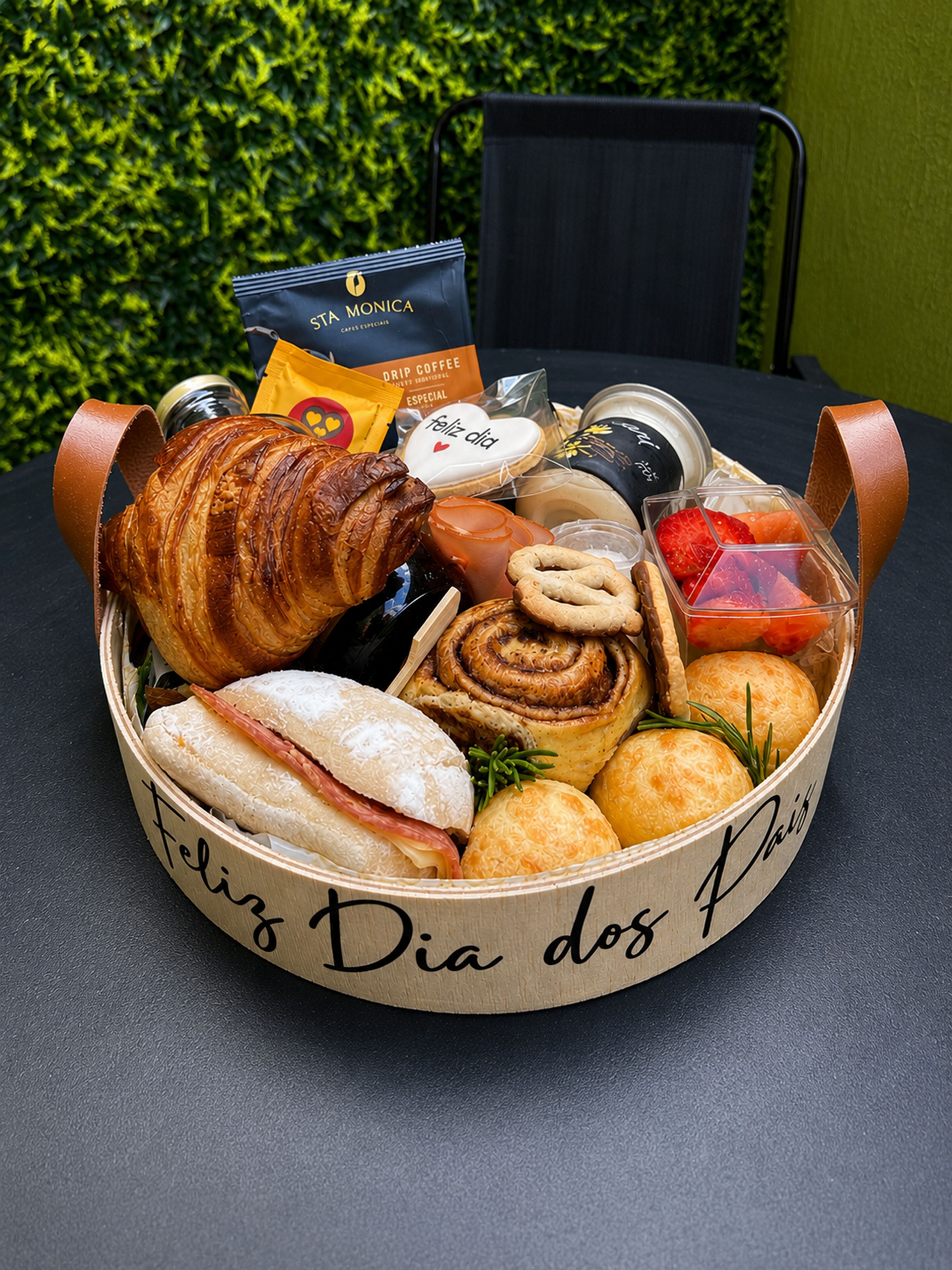 Uma cesta de madeira cheia de diversos doces e um saco de café, colocada sobre uma mesa com uma sebe verde ao fundo.