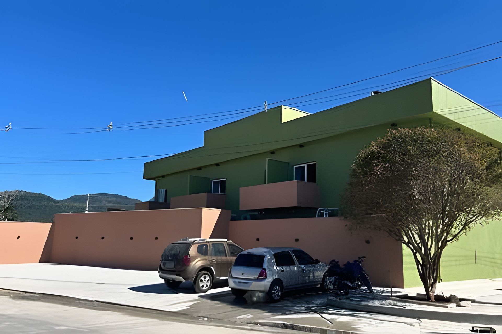 Un edificio moderno con techo plano, fachada verde y un área de estacionamiento con dos coches.