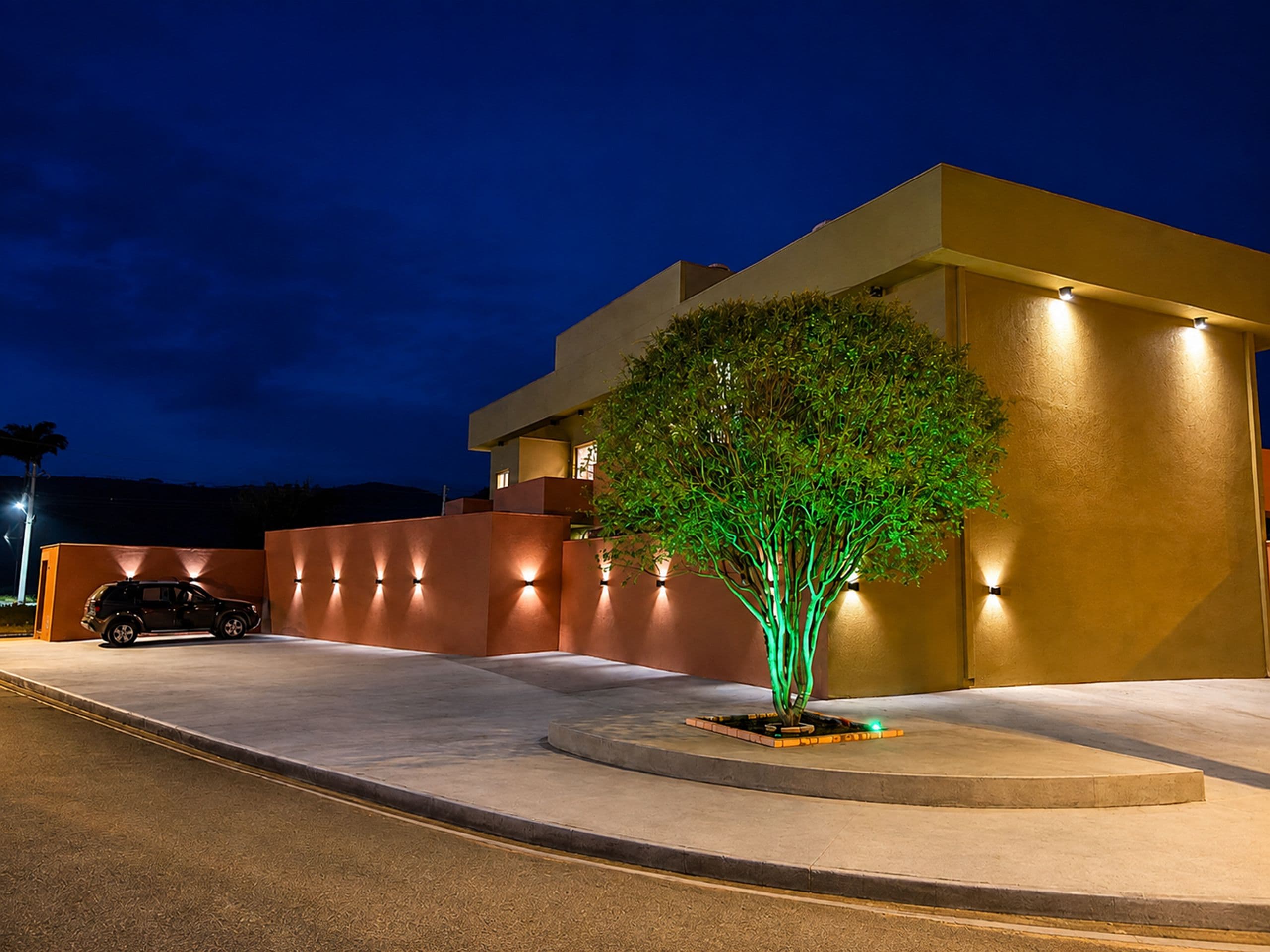 Un edificio moderno con un árbol iluminado por luces verdes, un coche estacionado al frente y una entrada bien iluminada.