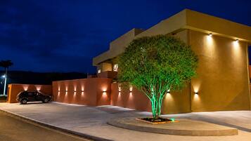 Un edificio moderno con un árbol iluminado por luces verdes, un coche estacionado al frente y una entrada bien iluminada.