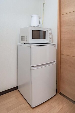 Un réfrigérateur blanc, avec un micro-ondes posé sur le dessus, un petit meuble blanc en dessous et une porte en bois sur la droite.