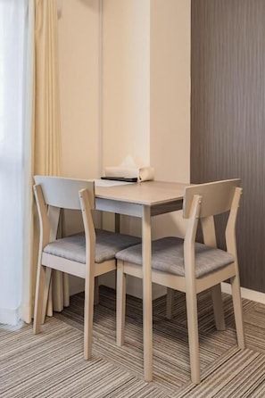 Ein Hotelzimmer mit einem Holztisch, vier Stühlen, einem kleinen Nachttisch und einem Fenster mit Vorhängen.