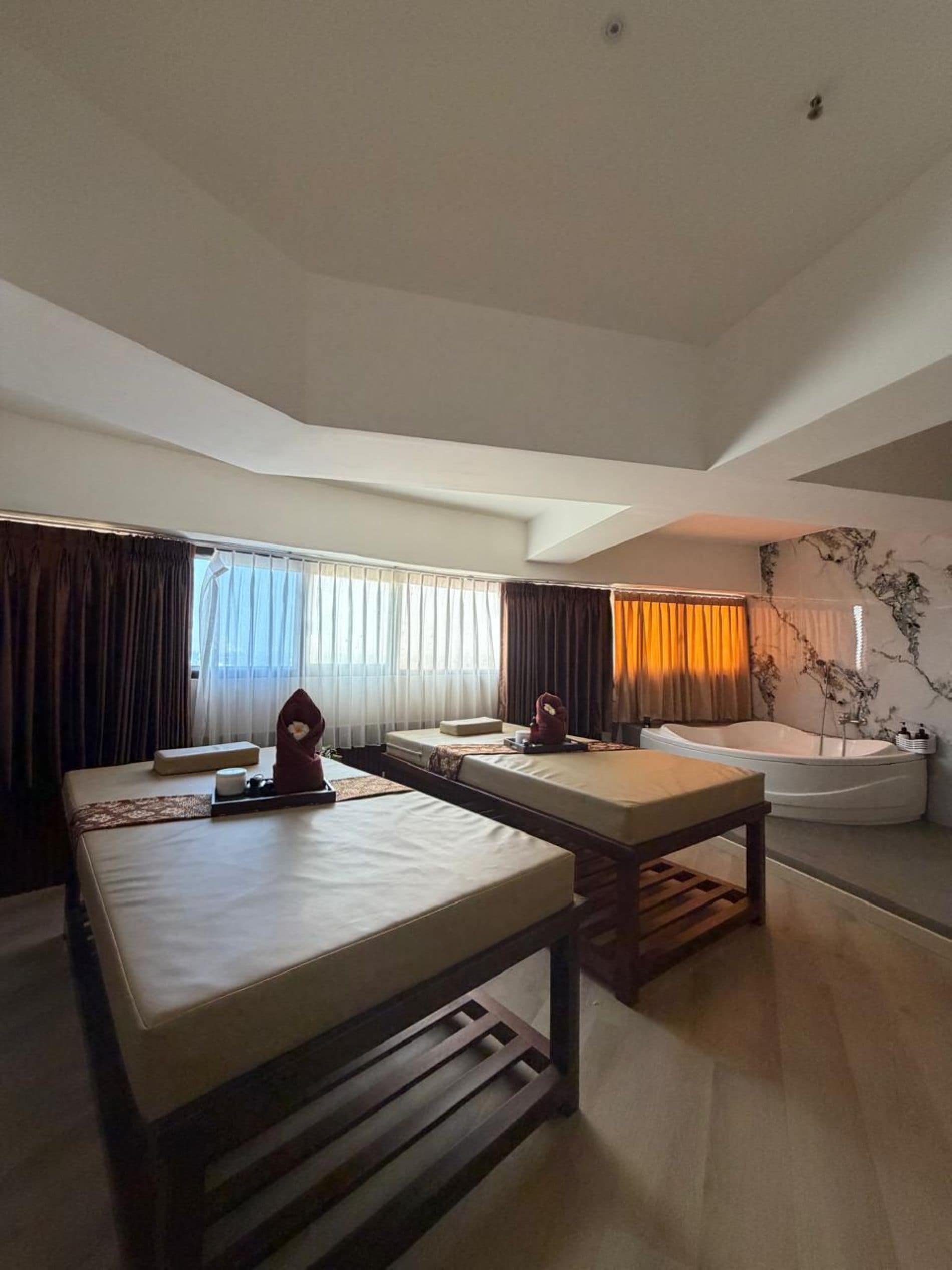A hotel room with a massage table, a separate bed, a bathtub, and a large window with curtains.