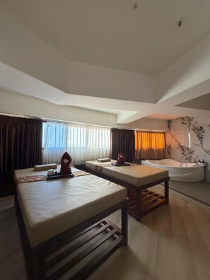 Habitación de hotel con una camilla de masaje, cama separada, bañera y un gran ventanal con cortinas.