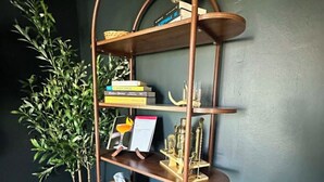 A brown, curved, multi-tiered wooden shelf with various items on it, including books, a glass, and decorative objects. - FOX DEN COTTAGE (Cleveland)