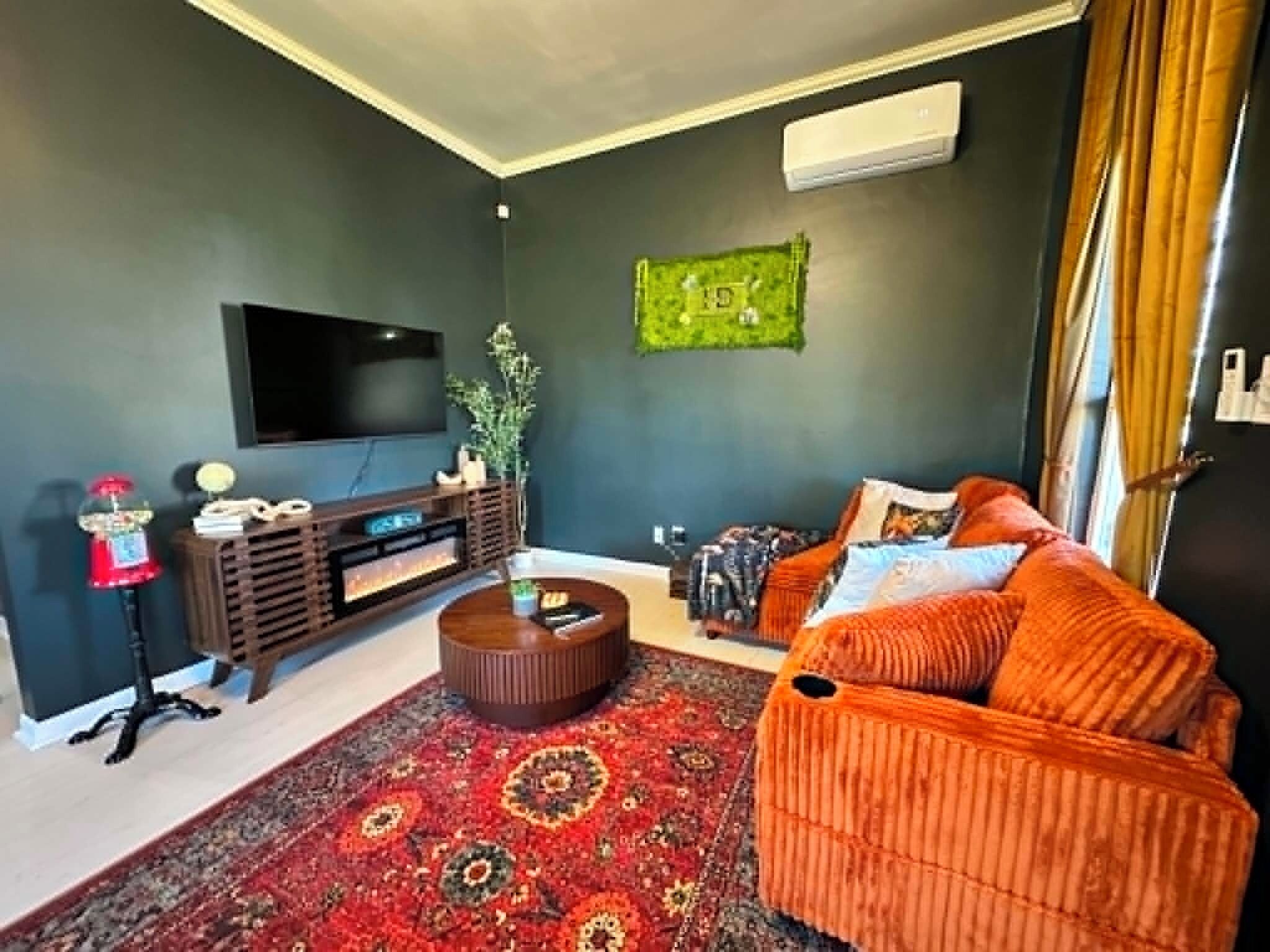 Una sala de estar con una televisión de pantalla plana, un mueble de entretenimiento de madera, un tapete estampado y un sofá naranja con cojines.