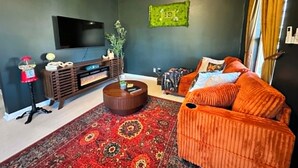 A living room with a flat-screen TV, a wooden entertainment unit, a patterned rug, and an orange sofa with cushions. - FOX DEN COTTAGE (Cleveland)